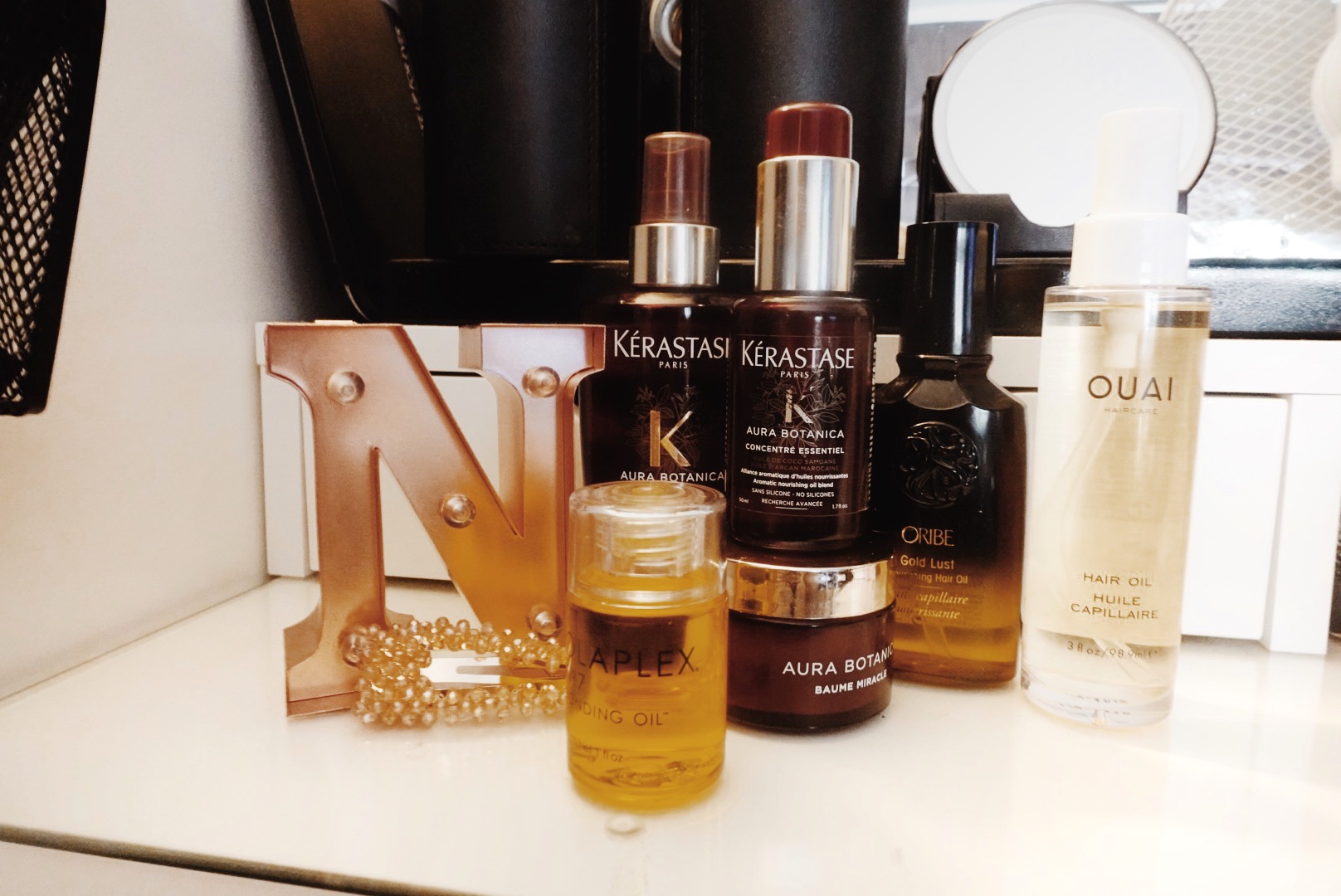 Bottles of various hair oils placed next to each other on the glass counter of a white vanity table. There’s also a rose gold coloured letter N used as decor along with the bottles of hair oil mentioned in the blog post.